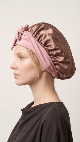 A woman wearing a silk sleep cap in two colors: a dusty pink headband with a large bow and adjustable elastic ties She is looking directly at the camera against a plain light background.