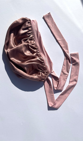 A dusty rose silk bonnet with a gathered elastic top and long matching ties, laid flat on a light gray background.